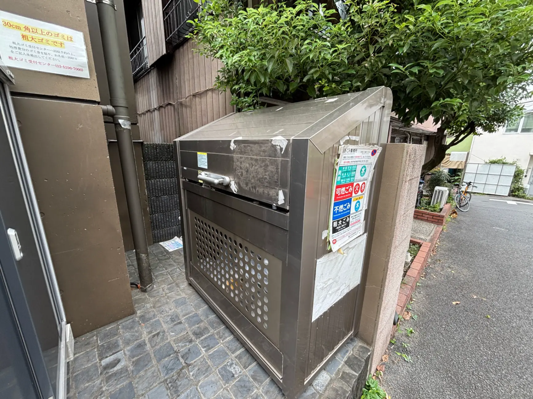 - 街中に設置された金属製のゴミ箱、周囲には樹木とコンクリートの壁、近くに自転車が置かれた静かな住宅街の風景
