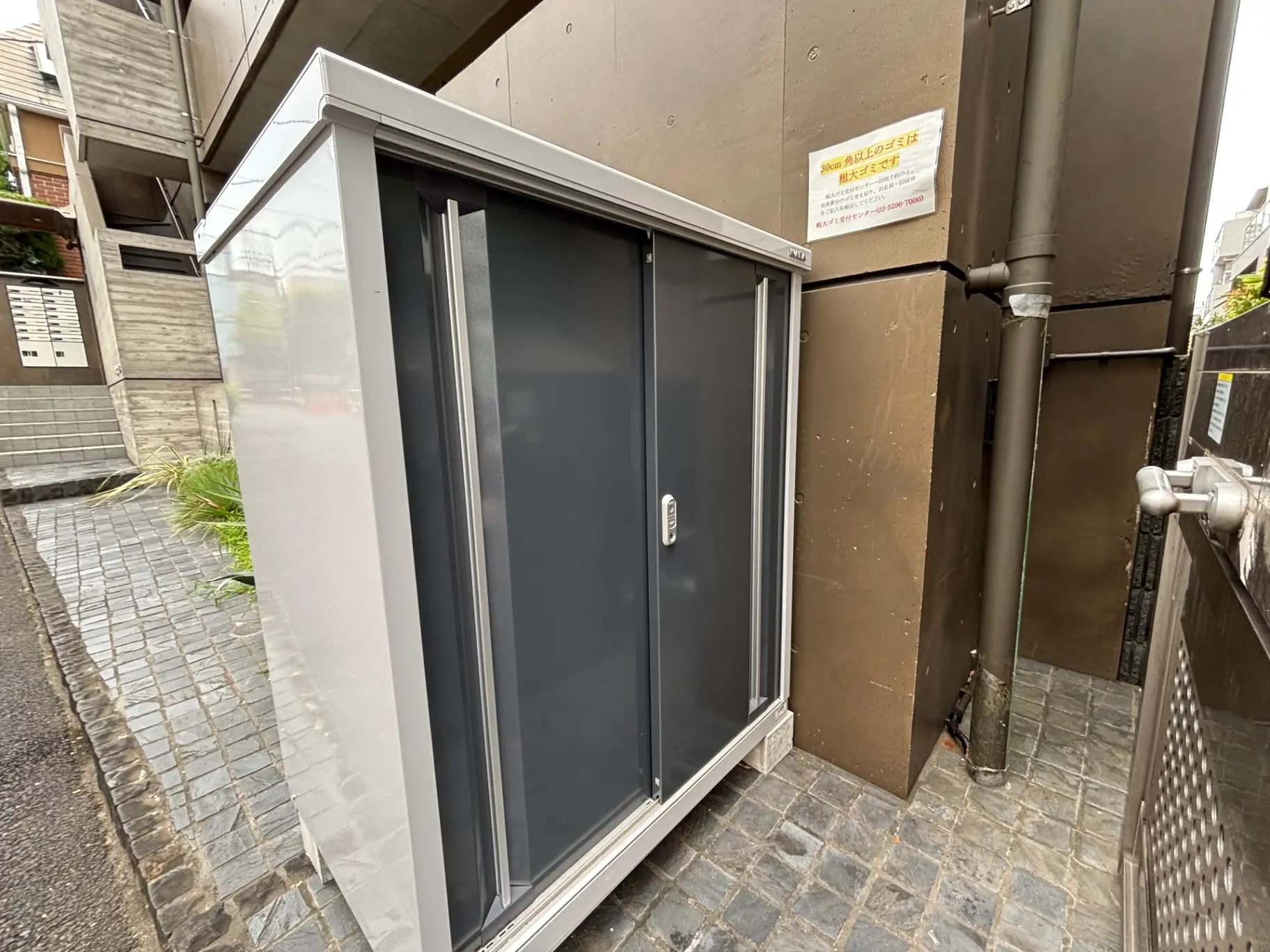 - モダンなデザインの集合住宅の側面に設置された自転車置き場、灰色の扉とポーラーホワイトの外観が特徴的な収納スペース