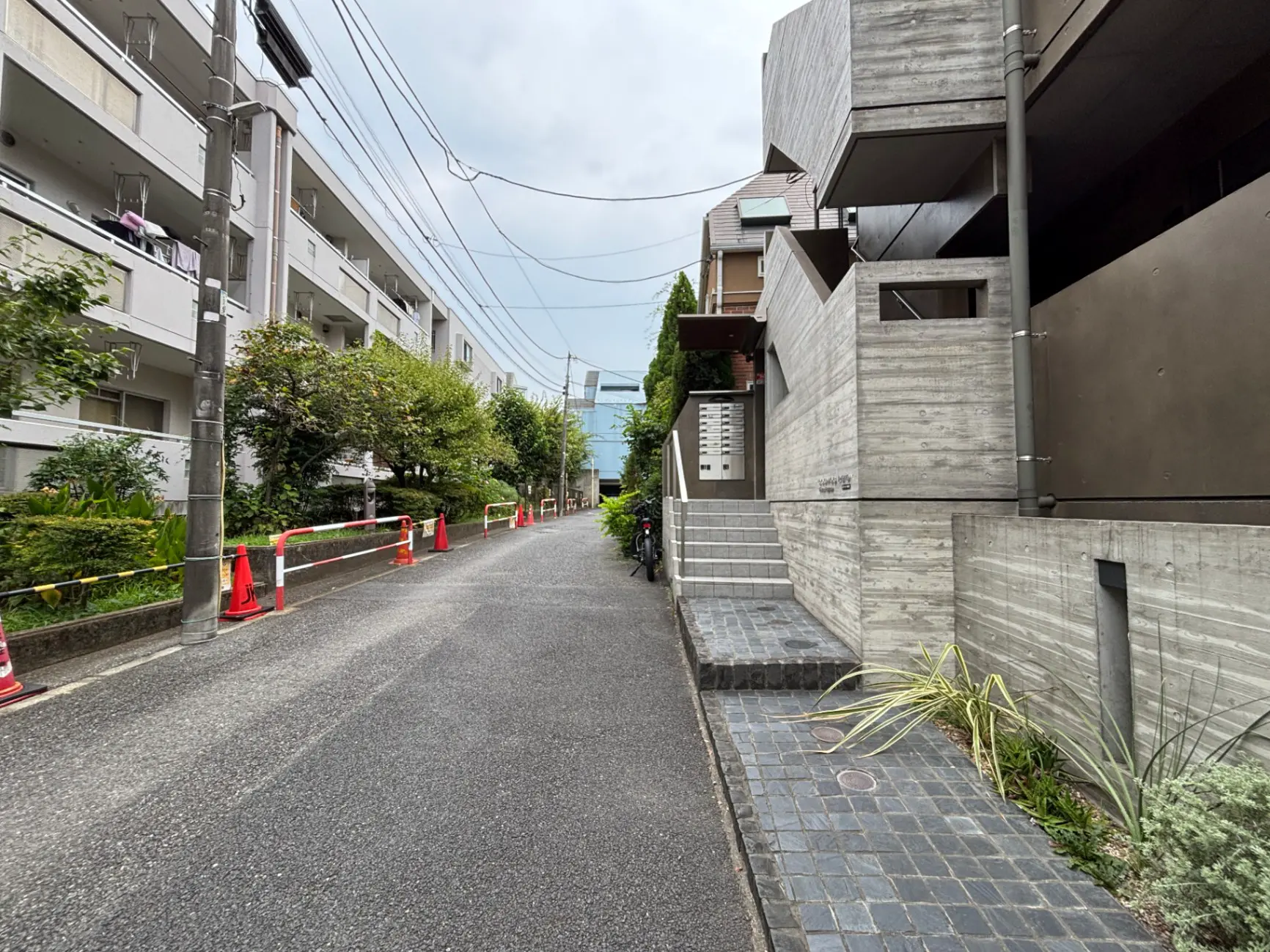 - 都市の景観を背景にした近代的なコンクリートとレンガ造りのアパートメント、植栽が施された歩道と交通標識が見える路地の風景