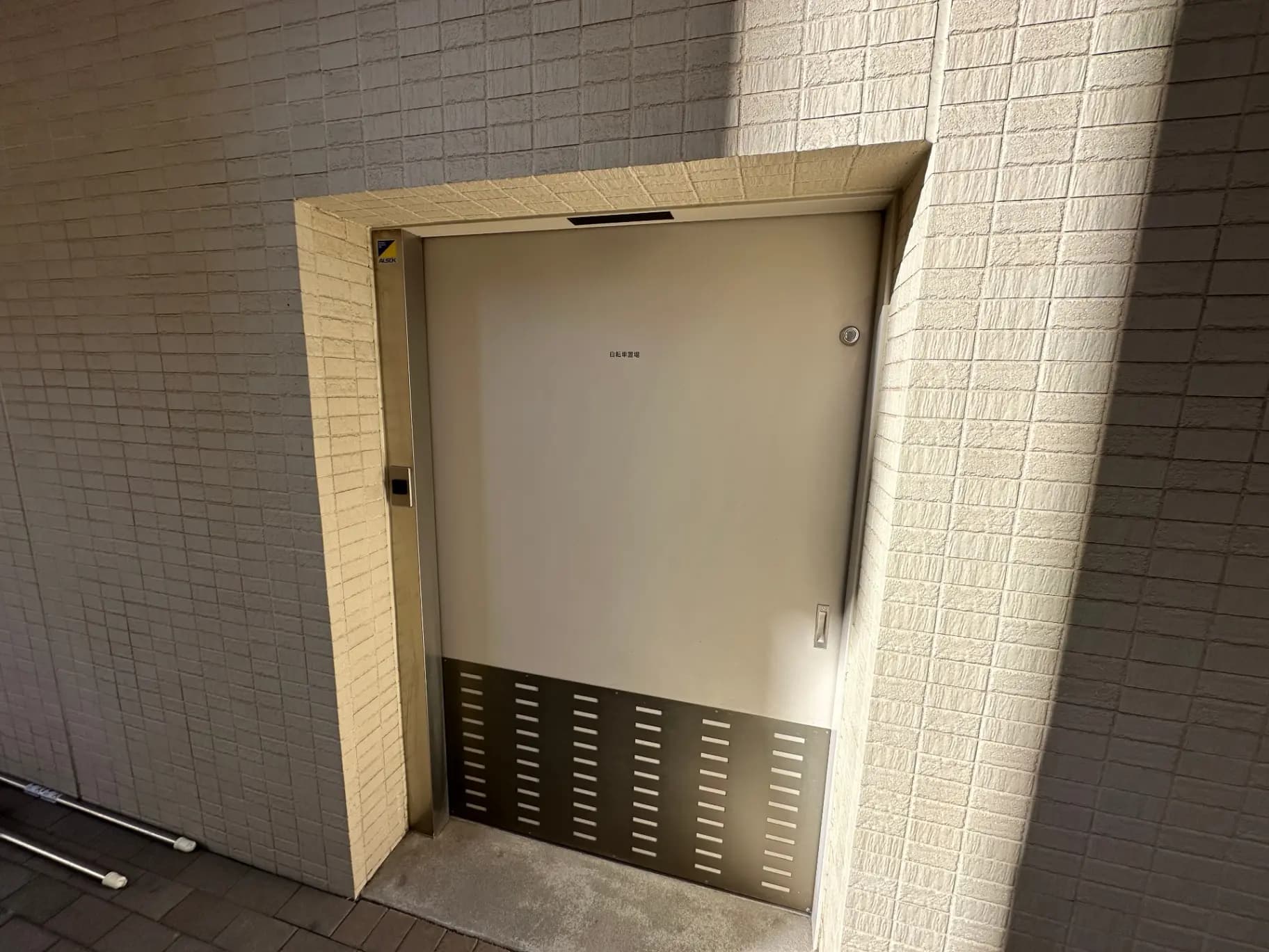 日差しが当たるモダンな外観の建物入口、ベージュの壁とシンプルなデザインのドア