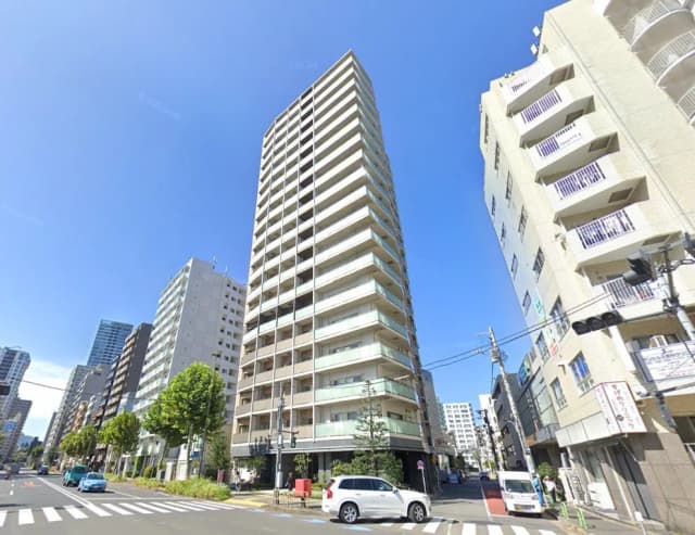 晴れた日の街角に立つ高層マンション、周囲には他のビルやアパート、横断歩道と走行する車が見える風景
