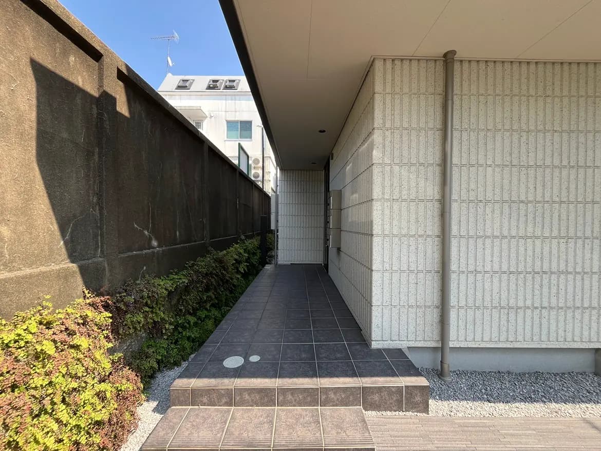 - モダンなエクステリアの入り口、タイル敷きの通路と緑の植栽が施されたアプローチ、青空の下にある建物の側面