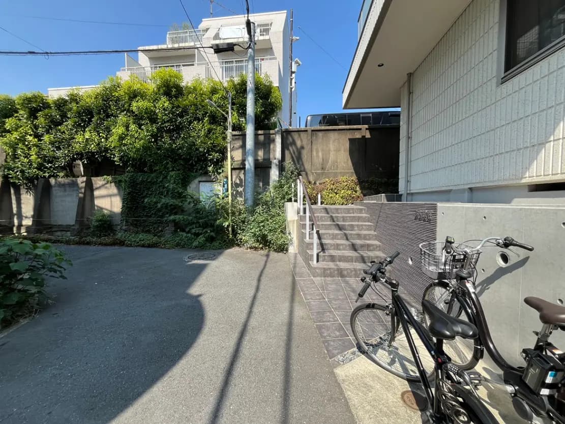 - 駐輪スペースに停められた自転車と、周囲の緑に囲まれた階段、青空が広がる住宅街の風景