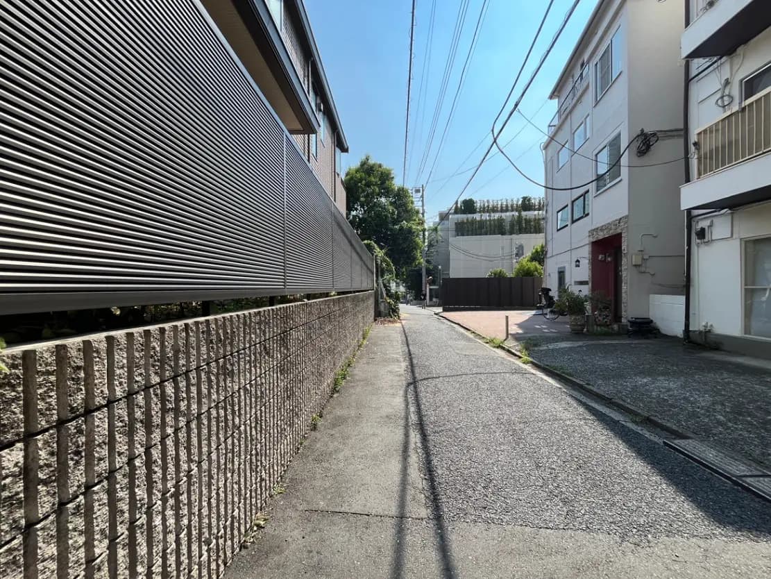 - 日中の静かな住宅街の狭い路地、左側にスラットフェンスがあり、右側には白い外壁の建物が並ぶ風景