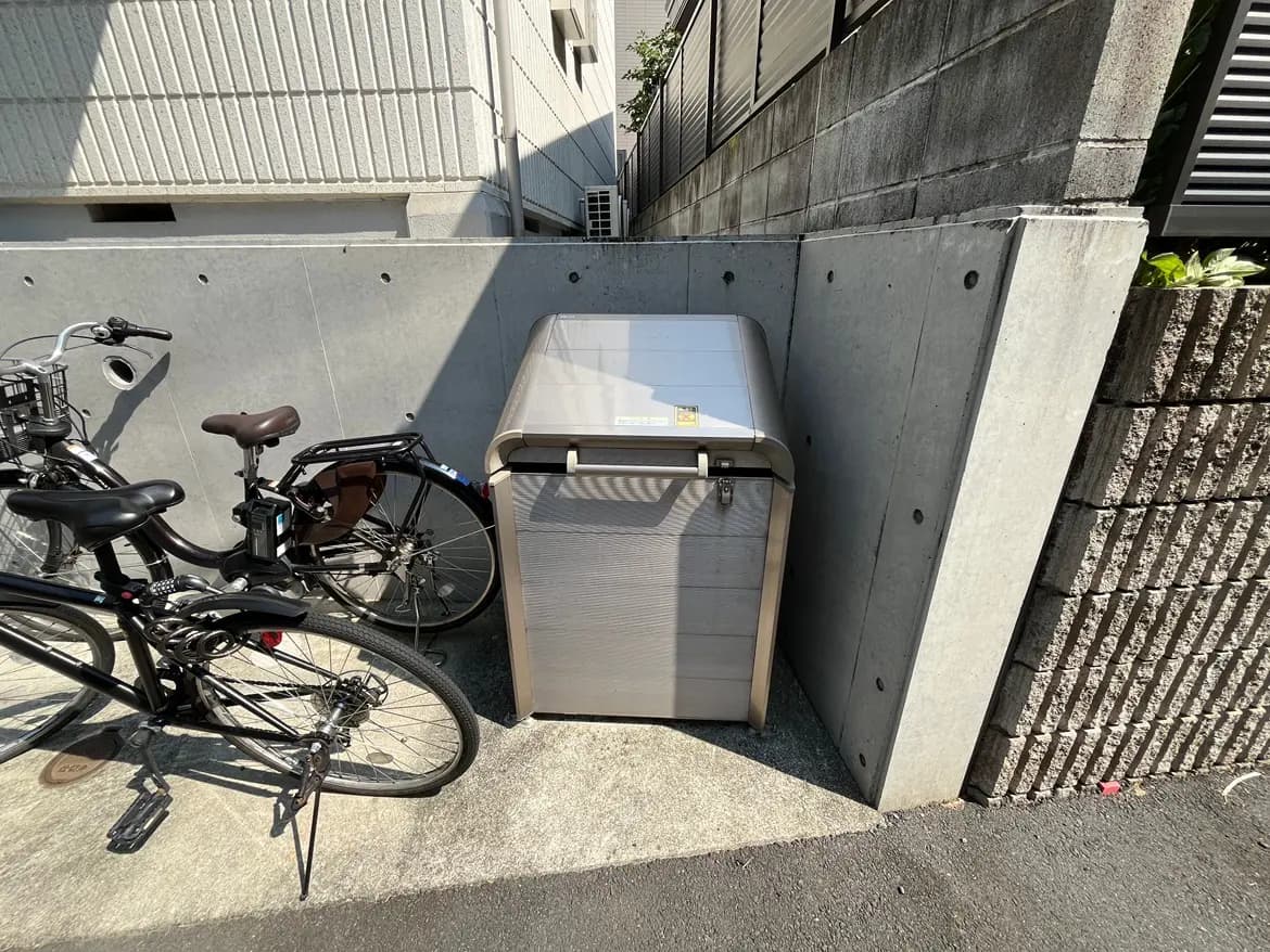 - 自転車が置かれたコンクリートの通路にある集合住宅用のゴミ捨て箱、周囲はブロック塀と壁に囲まれている