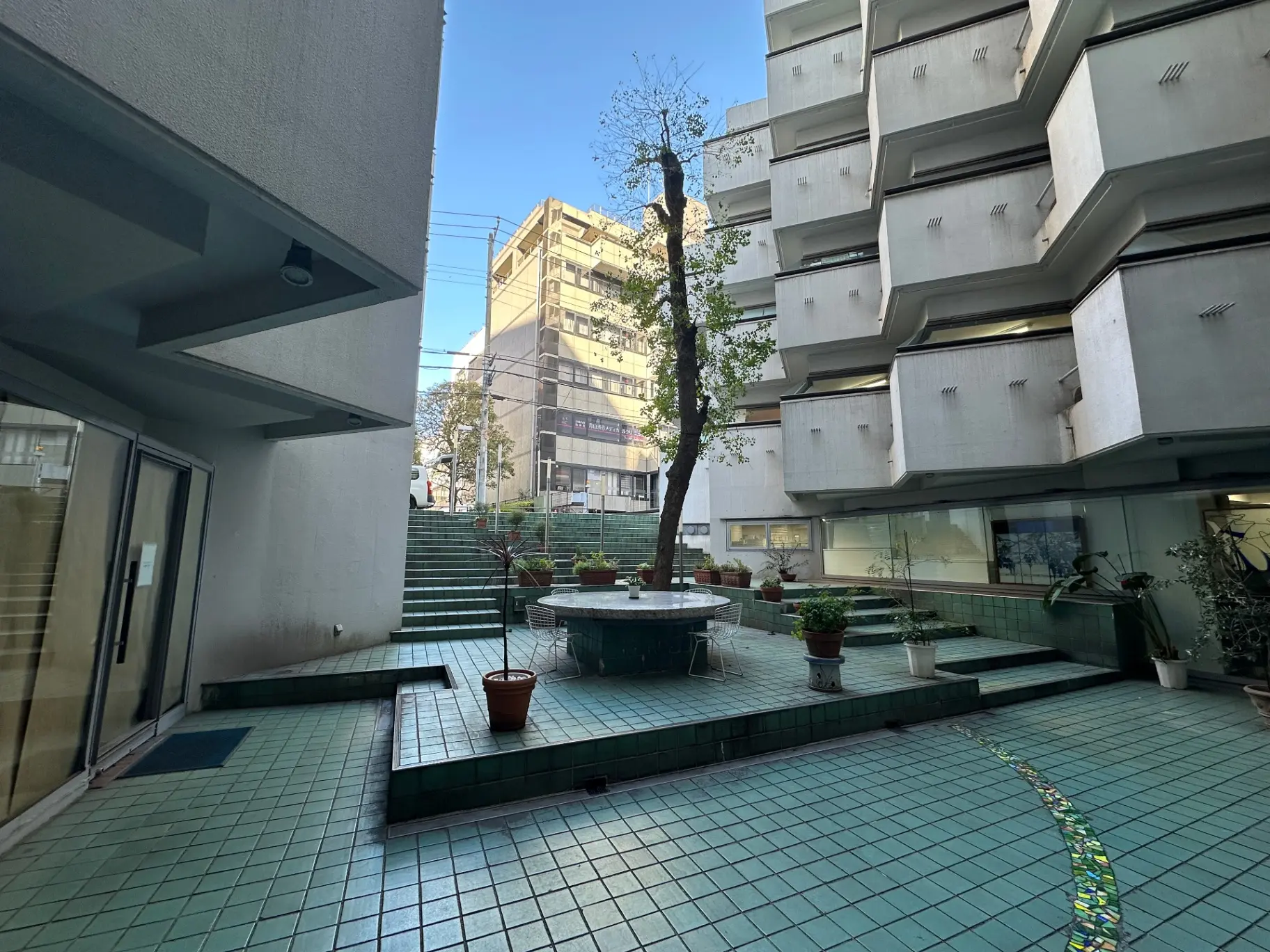 マンションの中庭、タイル敷きの空間、中央に噴水、背景に建物と樹木が見える都市風景