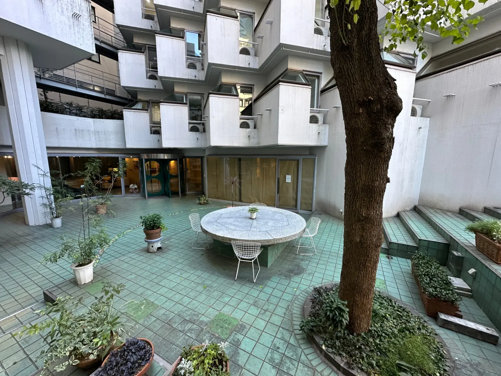 都会的なマンションの中庭、テーブルと椅子が置かれた緑のタイル張りのスペース、植物と木で装飾