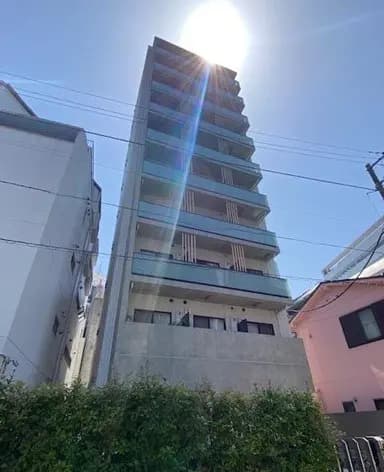 - 明るい青空の下、太陽光が差し込む新築の高層ビルの外観、左側には隣接する低層の建物が見える