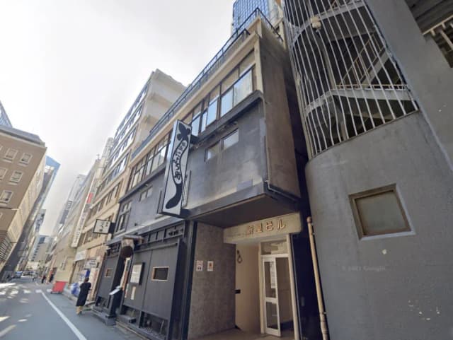 - 都会のビル群の中に位置する「新宿ビル」、黒い外観と特徴的な看板が印象的な建物の入口