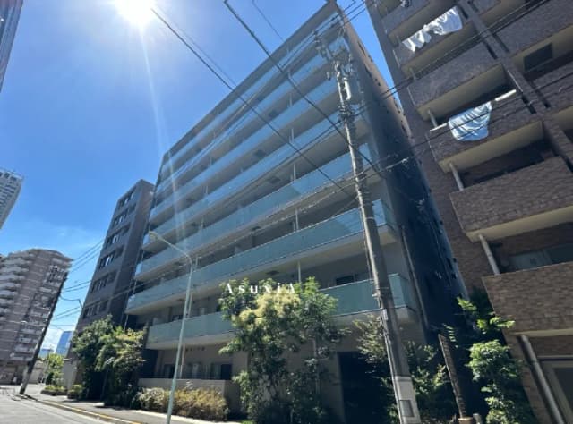 - 青空の下に立つ現代的なデザインのマンション、ガラスバルコニーと緑の植栽が特徴的な外観