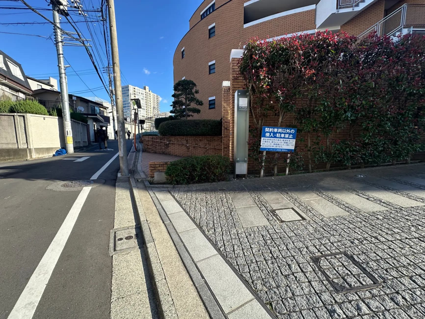 青空の下、閑静な住宅街の石畳の通りとレンガ造りのアパートメントの外観、電柱と植え込みが見える風景