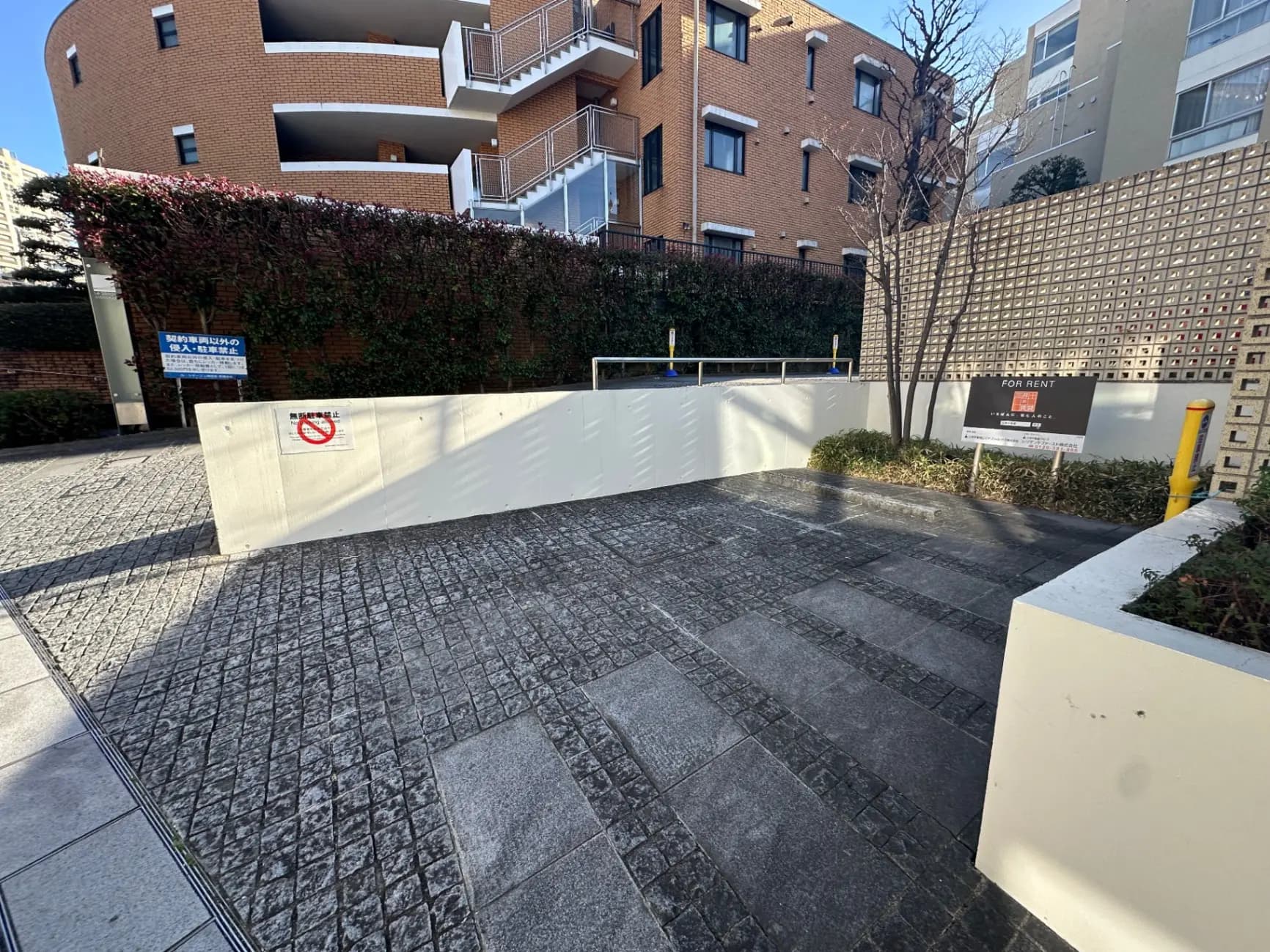 駐車場入口の横にある賃貸物件の看板、背景にはレンガ造りの建物と緑豊かな植栽