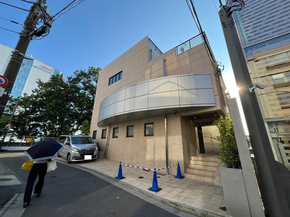 - 現代的なオフィスビルの外観、道路沿いに立つ、銀色のファサードと階段入り口、傘をさした歩行者が通り過ぎる風景