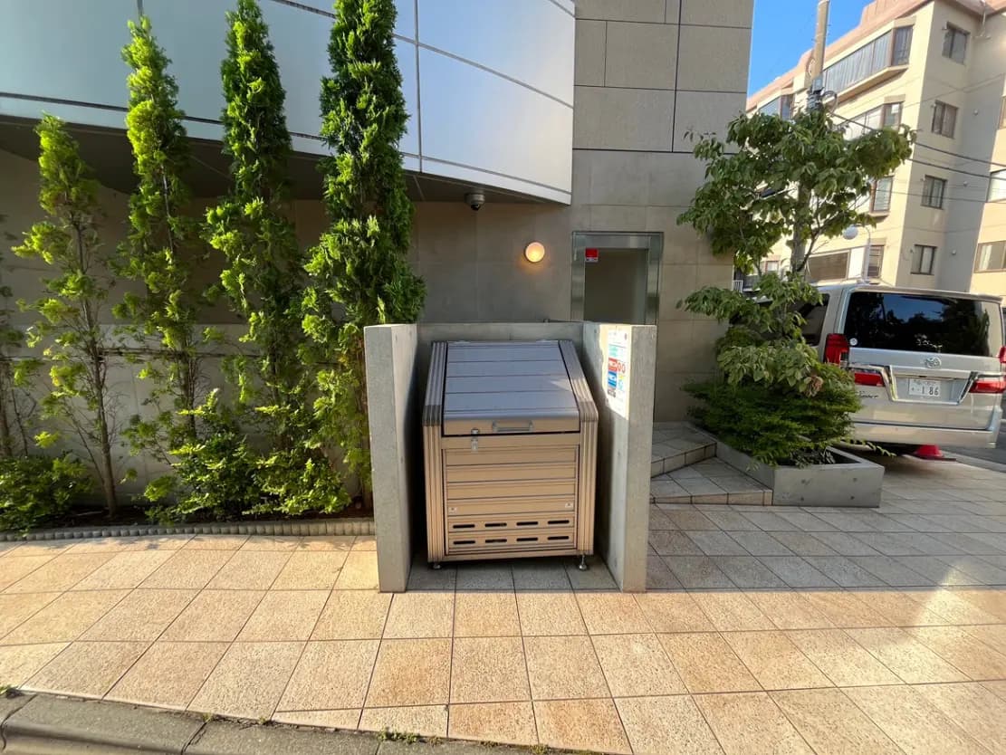 - 都市のエントランス近くに設置されたゴミ箱、周囲に緑の木々が植えられた整備された歩道の風景