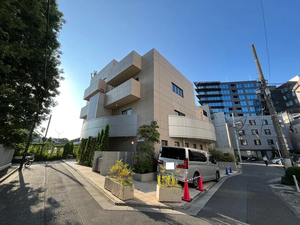 - モダンなデザインの建物、周囲に植栽と車両があり、青空が広がる街角の風景