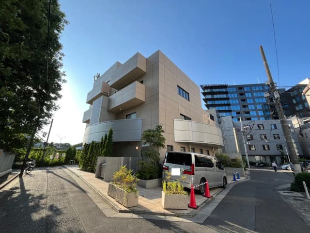 - モダンなデザインの建物、周囲に植栽と車両があり、青空が広がる街角の風景