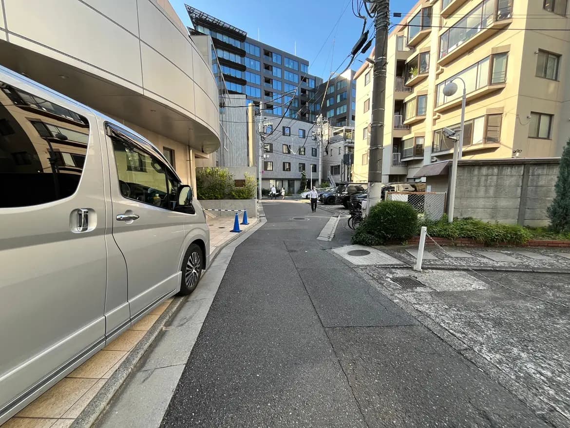 - 都市の住宅街の風景、銀色のバンが停車し、背後にオフィスビルやアパートが見える通りの一角
