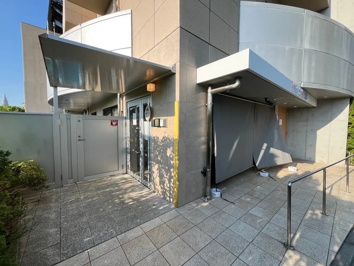- モダンな建物のエントランス、ガラスのフェンスを備えたアプローチ、屋根付きのエントランスと周囲に緑の植栽がある外観