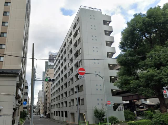 - 都市の中に立つ6階建てのアパートメントビル、周囲には高層ビルや緑豊かな木々、近くに交通信号と標識が見える通りの風景