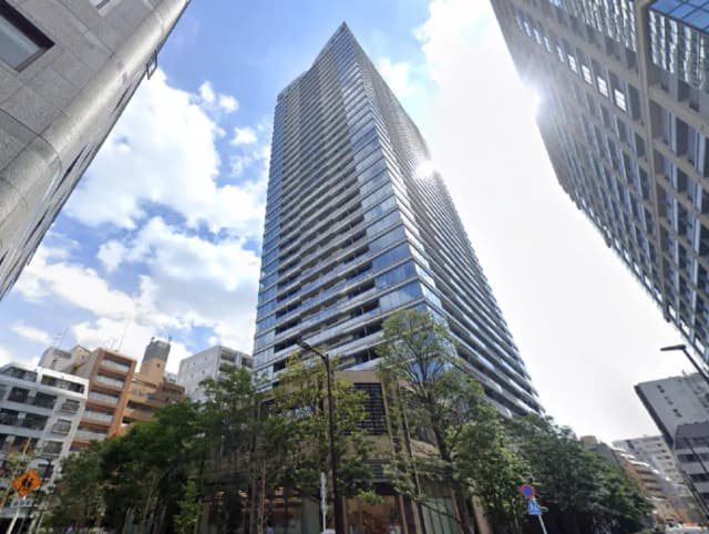 - 高層ビルが立ち並ぶ都市風景、青空と白い雲が広がり、手前には緑豊かな木々が生い茂る様子
