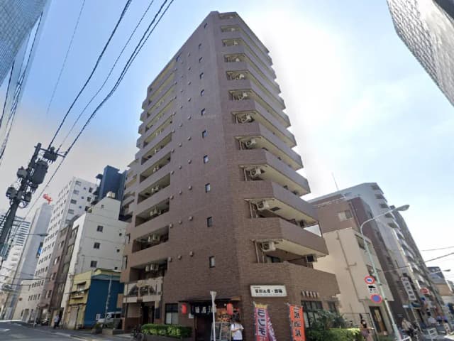 - 都市の真ん中に立つ高層マンション、レンガ調の外壁とバルコニーが特徴的な建物の外観、周囲には商業施設や電柱が見える街並み