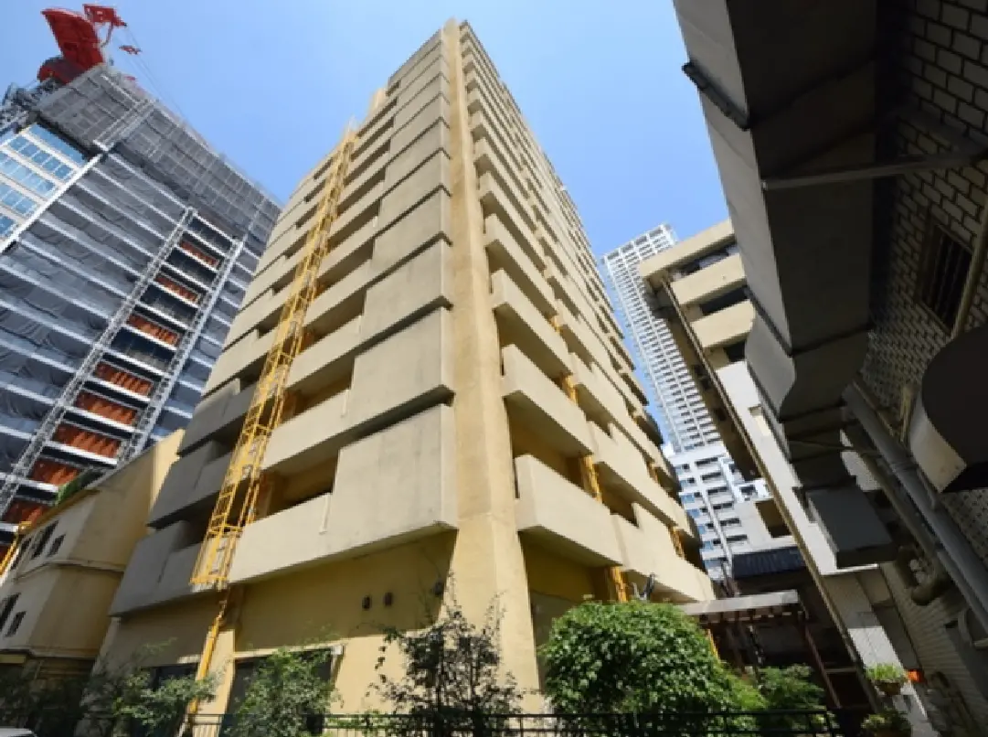 - 高層ビルの側面、周囲に新しい建物が並ぶ都市の景観、空を背景にしたコンクリートの外観と植栽のあるエリア