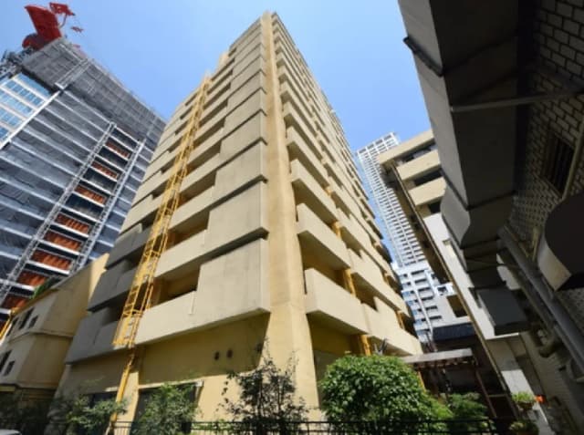 - 高層ビルの側面、周囲に新しい建物が並ぶ都市の景観、空を背景にしたコンクリートの外観と植栽のあるエリア