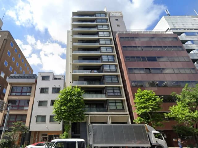- 都市の高層ビル街に立つ現代的なマンション、青空を背景にした外観と周囲の緑の木々が印象的な景観