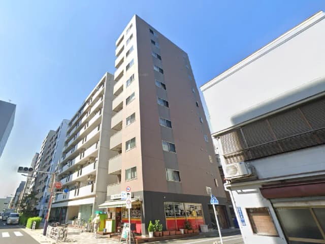 - 都市の一角に立つ現代的な多層マンション、晴れた空の下、周囲には店舗やカフェが点在する風景