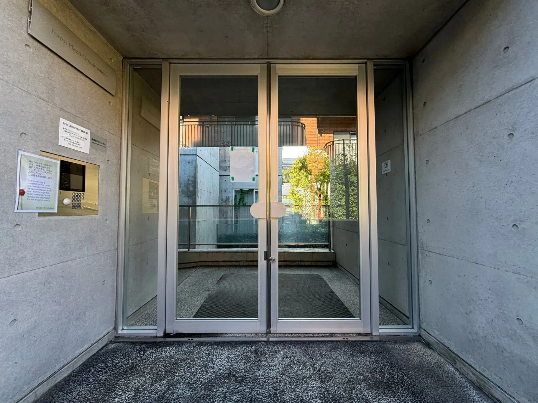 モダンな建物のエントリーウェイ、インターホンと案内掲示板があり、ガラス扉を通して外の緑が見える光景