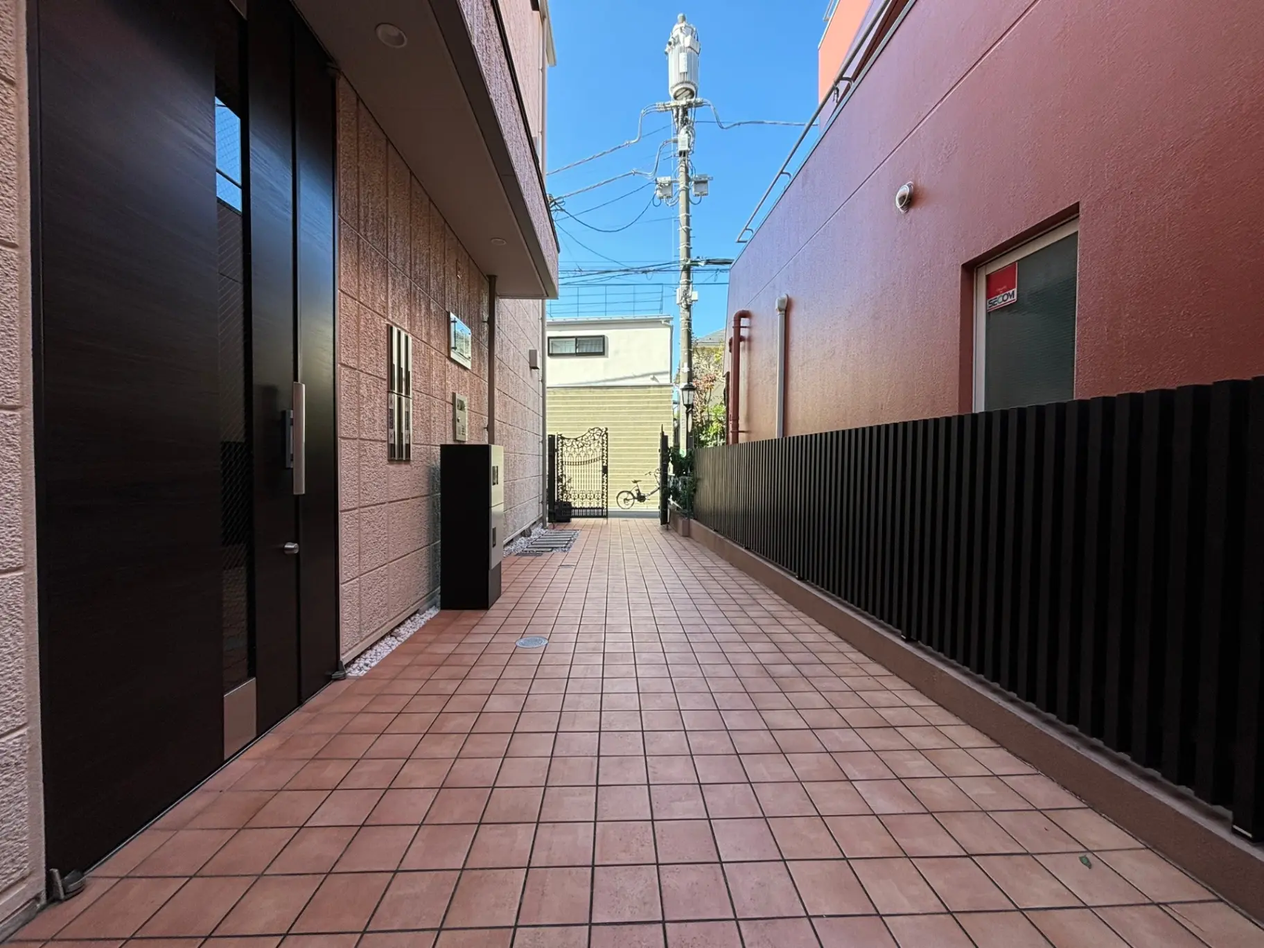 都市のアパートビルの間の狭い通路、整然と並んだドアとフェンス、明るい日差しが降り注ぐ舗装された歩道