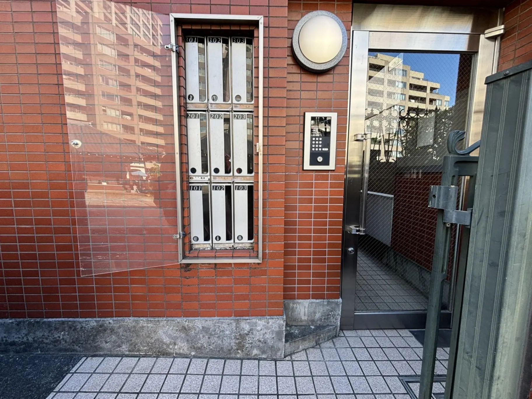 赤いタイル張りの壁に取り付けられた郵便受けとインターホン、都市の建物が反射するガラスのドア