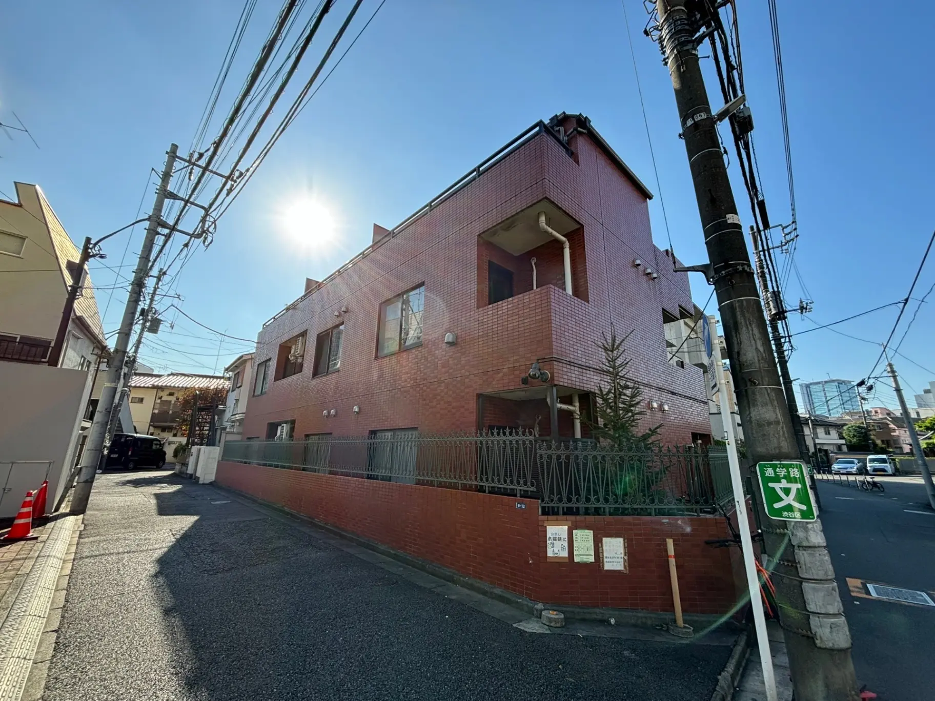 青空と日差しが照らす街角のピンク色のアパート建物、電柱や電線が交差する都市風景