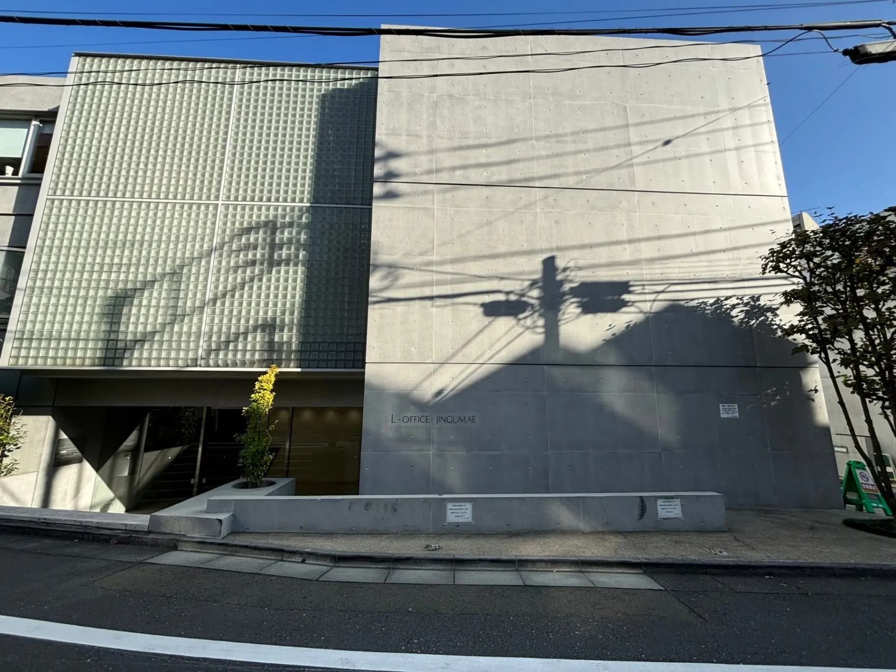 現代的なオフィスビルの外観、コンクリート壁に影が落ちるデザイン、街路樹とクリアな青空