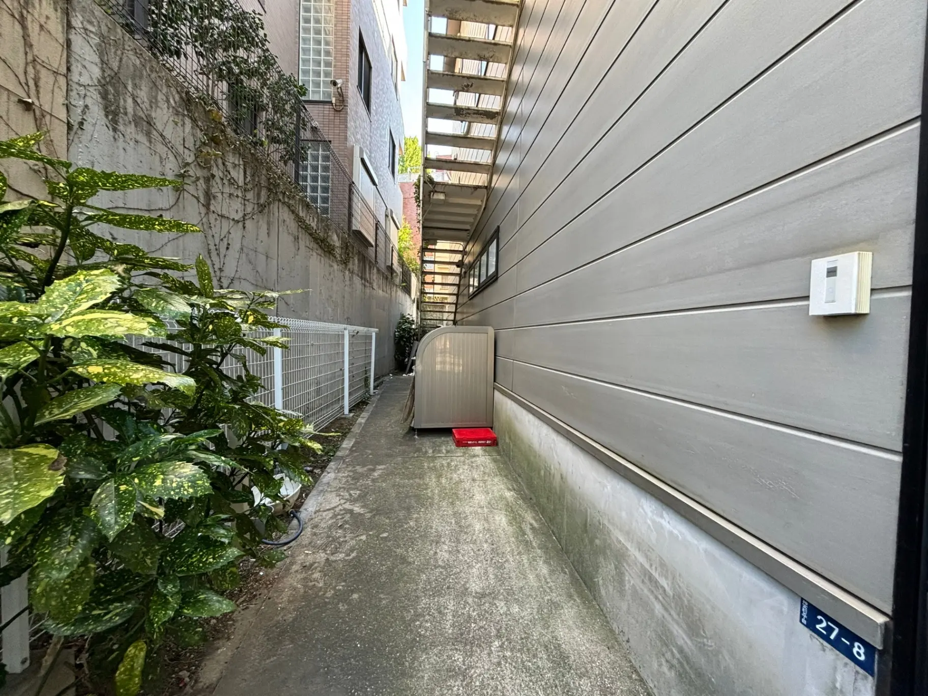 狭い都市の路地、左側に緑の植物、右側には現代的な建物の壁と窓、直射日光が差し込む明るい景色
