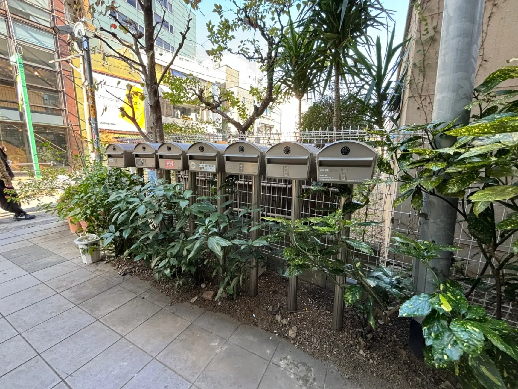 都会の歩道に並んだ複数の郵便受け、緑豊かな植栽と明るい日差しが特徴的な街並み