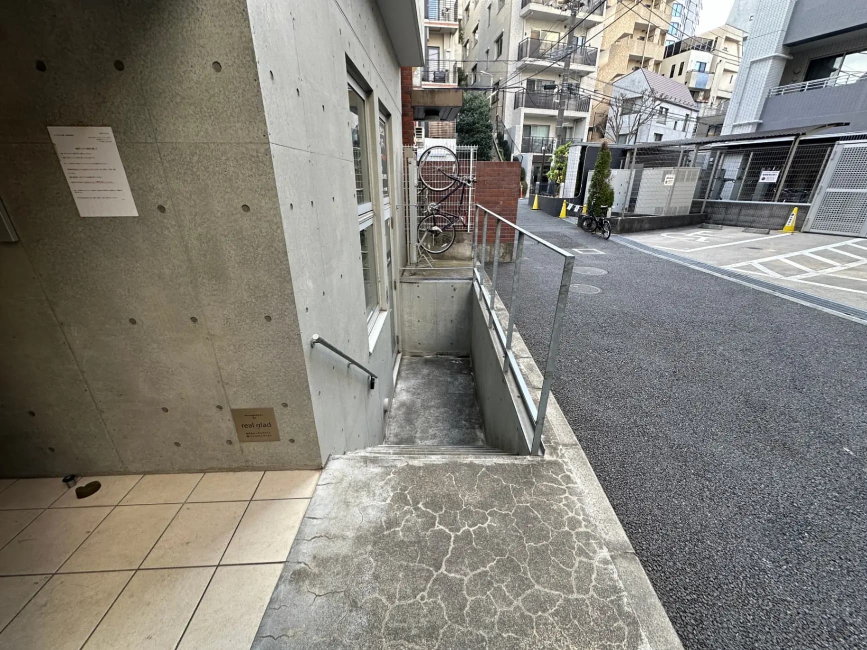 都市部のアパート外観、コンクリートの壁と地下への階段、路上駐輪スペース