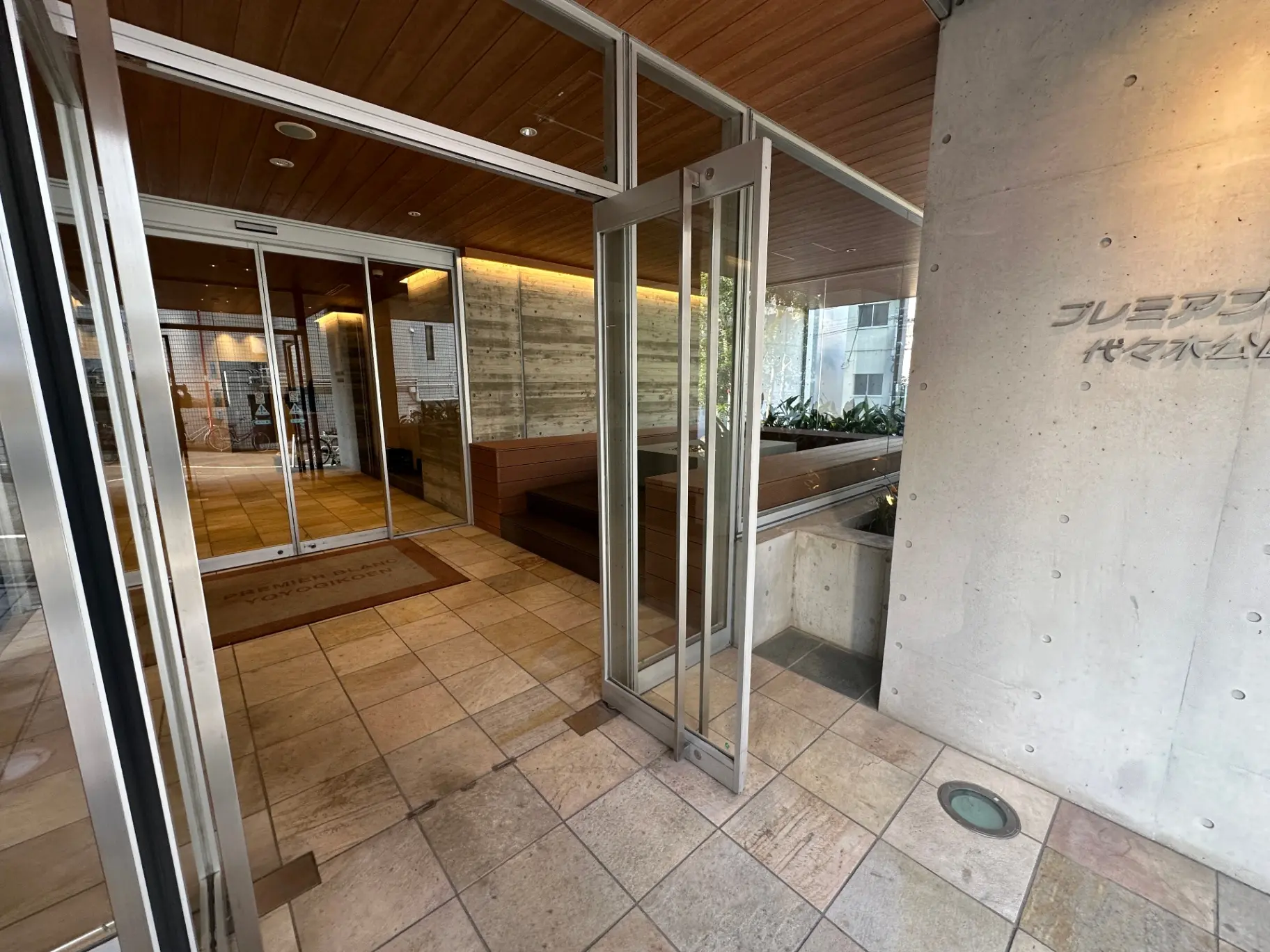 モダンなエントランスの自動ガラスドア、木目調の天井、コンクリートの壁装飾がアクセントの建物入り口