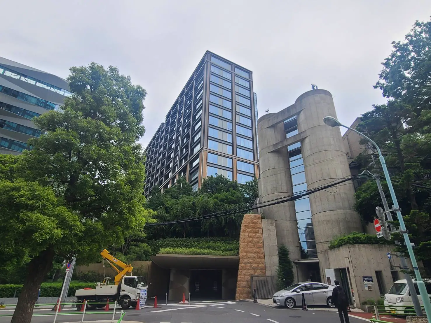 - 現代的なデザインの高層ビルとコンクリート構造の建物、周囲に緑豊かな木々と工事車両が見える街の風景