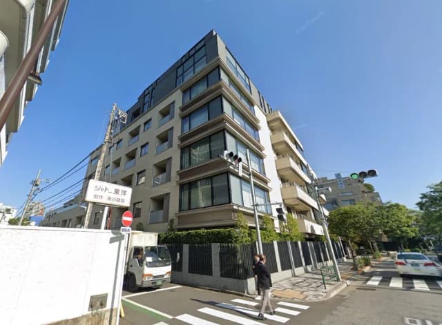 都市の交差点にあるモダンなマンションの外観、日差しの中で歩行者が横断歩道を渡る光景