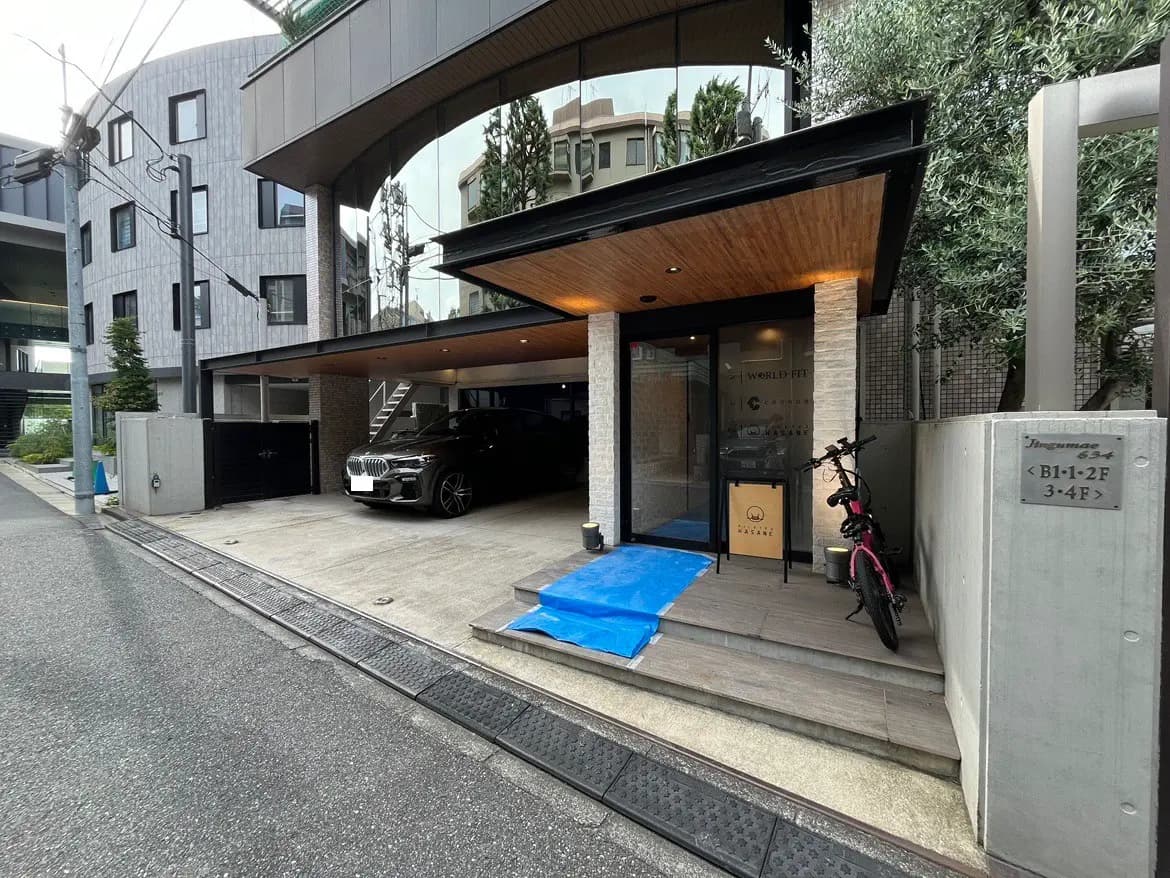 - モダンなデザインの建物のエントランス、黒い車と自転車、木製のデッキと玄関前の仕上げ、周囲の緑が特徴的な外観