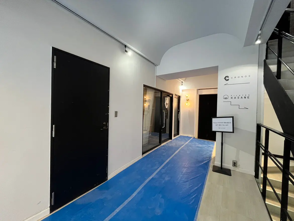 - シンプルでモダンなエントランスホール、黒いドアと白い壁、青いシートが敷かれた通路、奥に見える照明のある空間