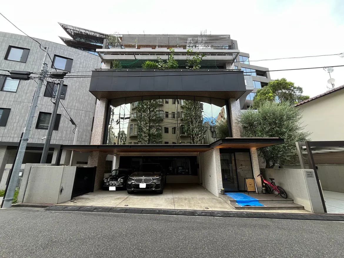 モダンなデザインの住宅外観、ガラス張りのファサード、駐車場には3台の車、周囲に緑の植栽があるエントランスエリア