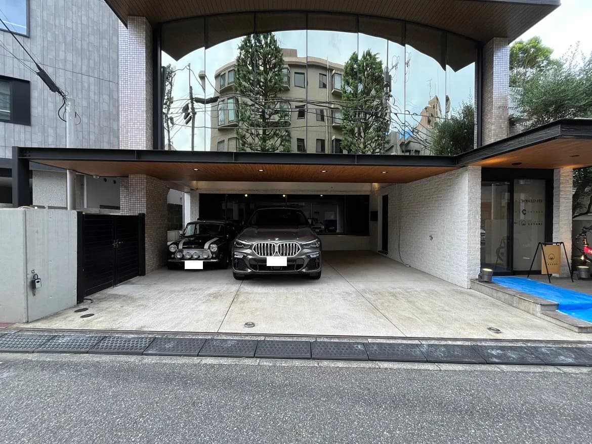 - モダンな外観の建物前に広々とした駐車スペース、2台の車が停まっている様子、ガラス張りのエントランスと木製の屋根のデザイン