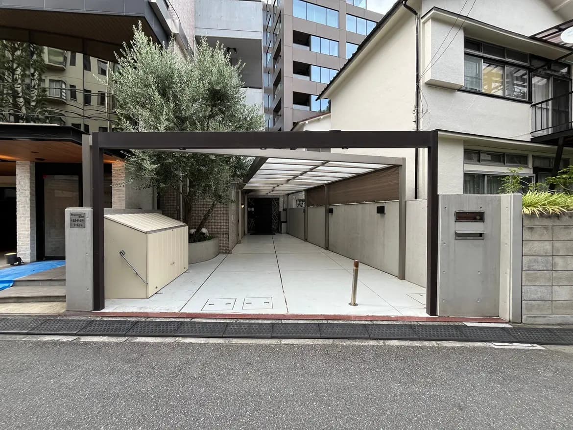 - モダンなデザインの住宅入り口、ガラス張りの外壁と明るい歩道、都市の景観を背景にした緑の植物が配置された駐車場エリア