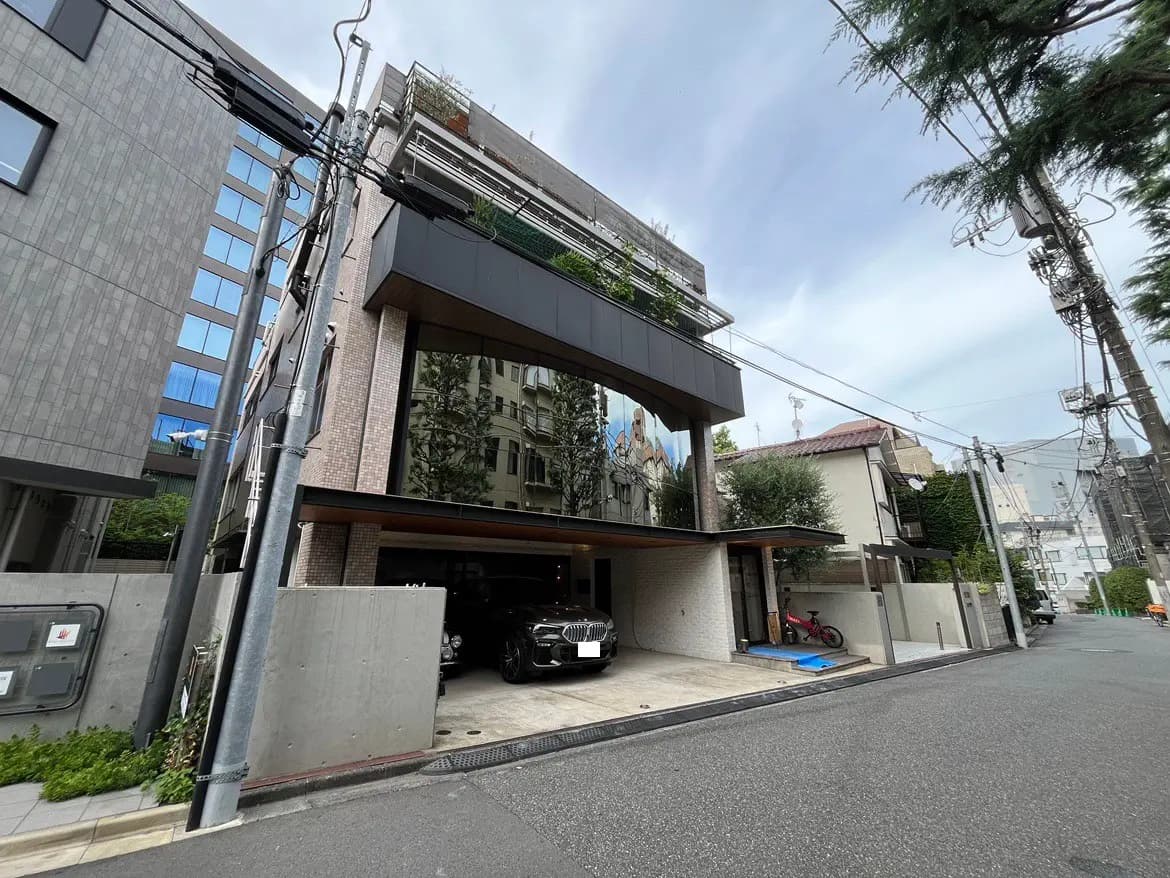 - モダンなデザインの住宅、隣接する建物と電柱がある街並み、反射するガラスのファサードと緑の植栽が特徴的な外観