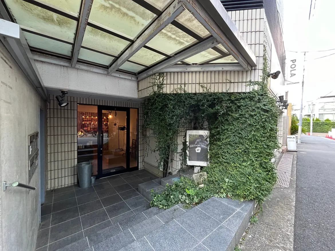 - 自然のグリーンが彩るエントランス、モダンなデザインの建物入り口と屋根付きのアプローチ、ガラス扉からの明かりがもれる様子