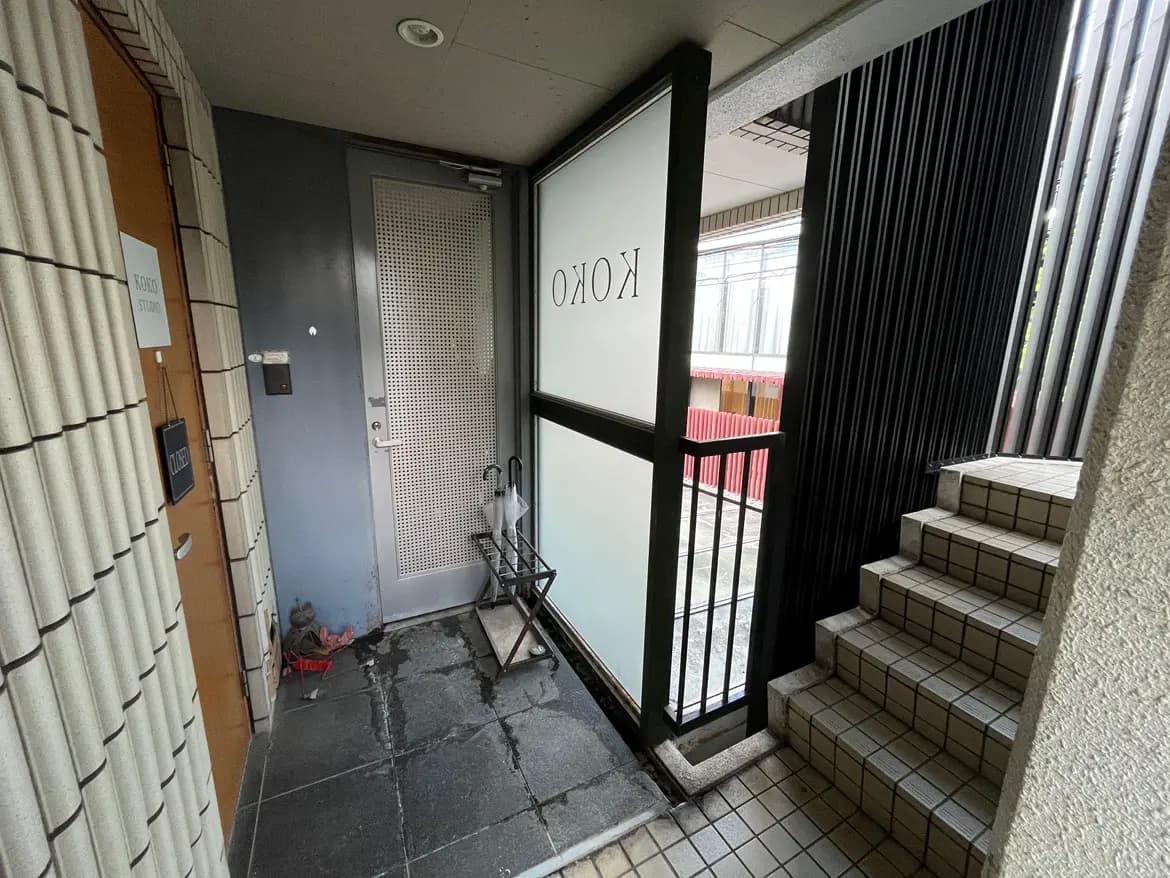 - モダンなデザインのエントランス、ガラスのドアから自然光が差し込み、左側には階段とシンプルなインテリアが配置された空間