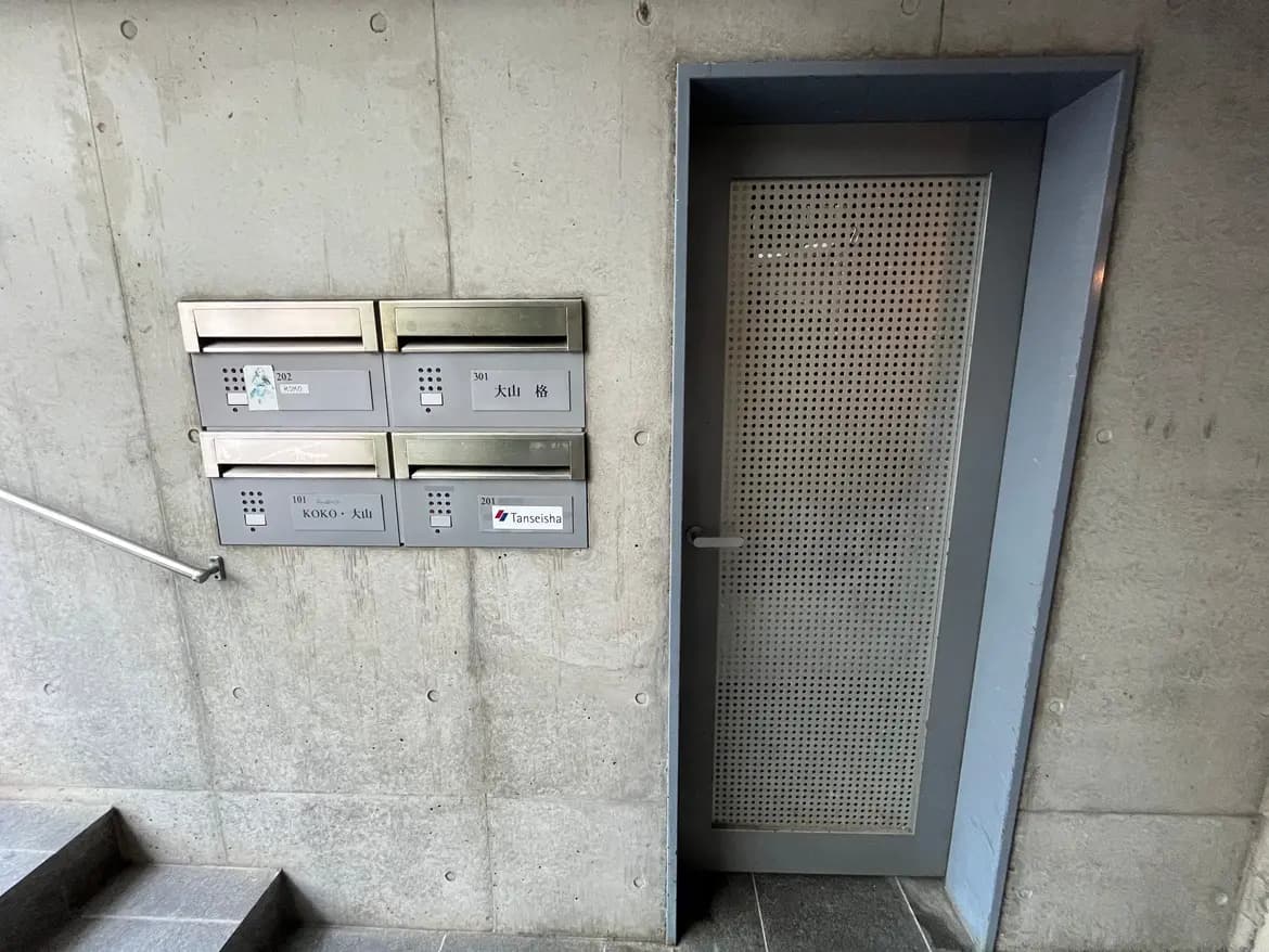 - コンクリートの壁に取り付けられた郵便受け、横にある個別のドア、階段と手すりのあるエントランスの風景