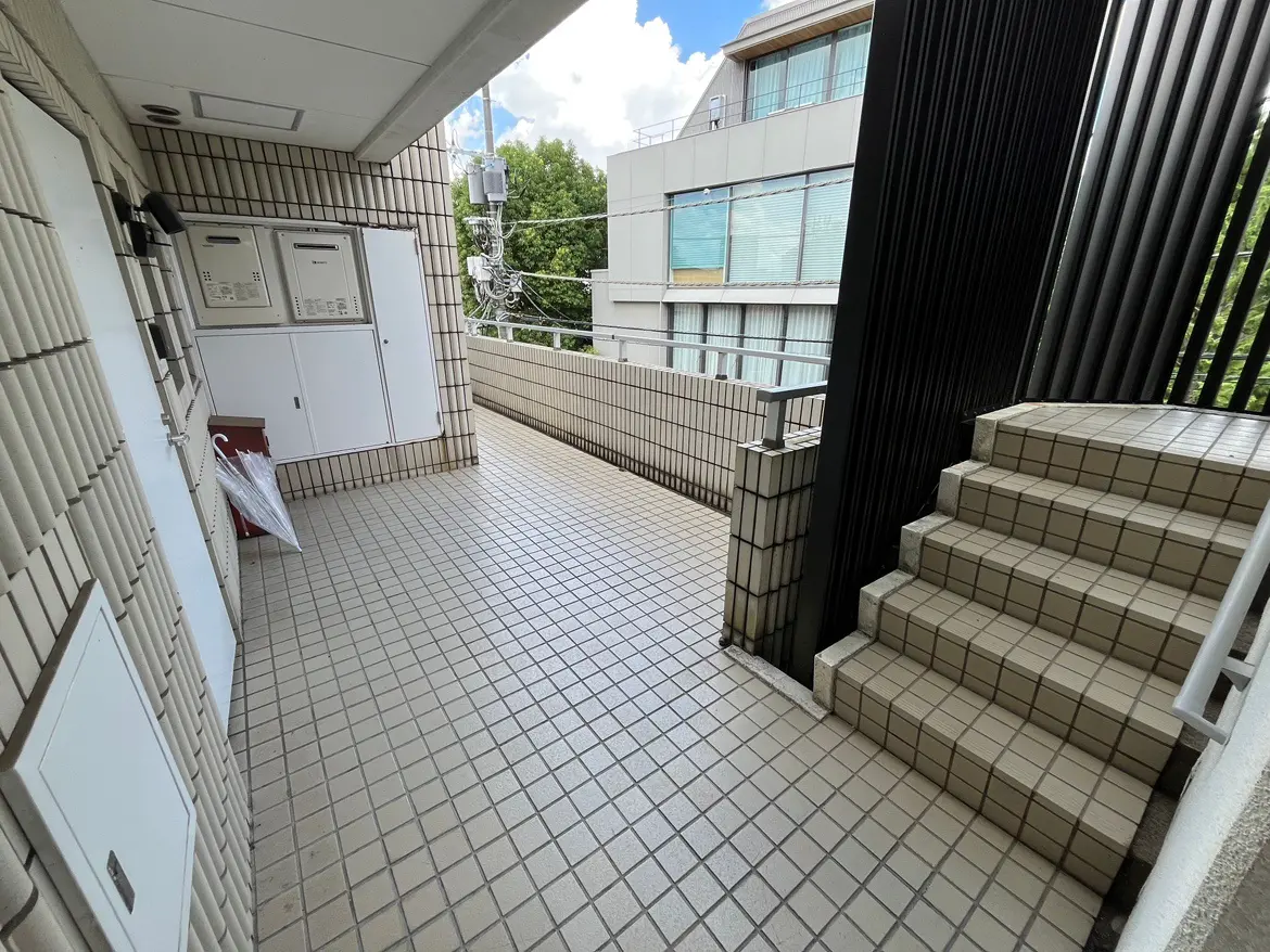 - 明るい屋外の通路、タイル張りの床と階段、郵便受けが設置された壁、周囲の緑が見える景観